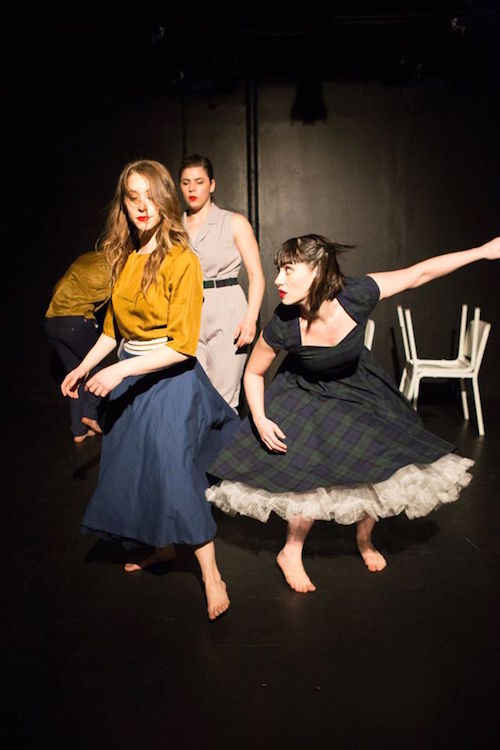 A woman in a plaid dress with a petticoat peaking out from underneath crouches and peers up at a woman in a yellow blouse and blue skirts